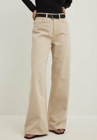 Jeans beige a gamba larga realizzati in tessuto resistente, dotati di vita alta, passanti per cintura, tasche frontali e orlo dritto. Cintura nera inclusa.