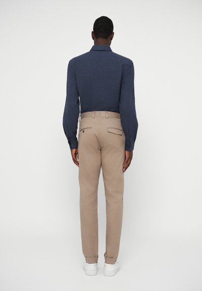 Jūras spēku pogu krekls ar garām piedurknēm, savienots ar bēšām biksēm ar aizmugurējām kabatām un baltām sporta apavām, skatījumā no aizmugures.