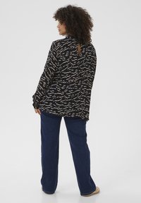 Chemise noire avec un motif beige ondulé, présentant un design en trou de serrure à l'arrière. Assortie à un pantalon large bleu foncé et à des chaussures de couleur claire.