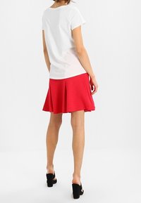 Biała koszulka z krótkim rękawem w połączeniu z czerwoną spódnicą rozkloszowaną do kolan. Modelka nosi czarne sandały na wysokim obcasie. Teksturowana tkanina.