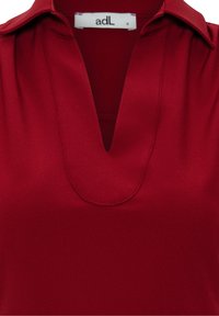Blouse rouge avec un col en V et un col, montrant une étiquette blanche avec le nom de la marque "adL" à l'intérieur de la zone du col.