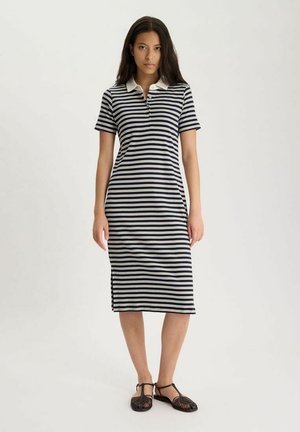 Femme portant une robe polo rayée horizontale noire et blanche à manches courtes et des sandales tissées noires, debout devant un fond clair.