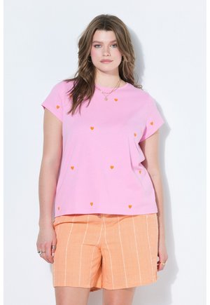 Junge Frau mit welligem braunem Haar, die ein pinkes Shirt mit kleinen orangen Herzen und orange gestreifte Shorts vor weißem Hintergrund trägt.