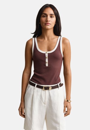 Femme portant un débardeur marron sans manches avec des bordures blanches et des boutons, associé à un short blanc taille haute et une ceinture marron foncé.