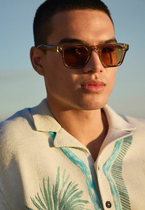 Mladý muž nosiaci slnečné okuliare s jantárovým zafarbením a krémový pletený sveter s modrými a zelenými tropickými listovými vzormi, vonku v jemnom dennom svetle.