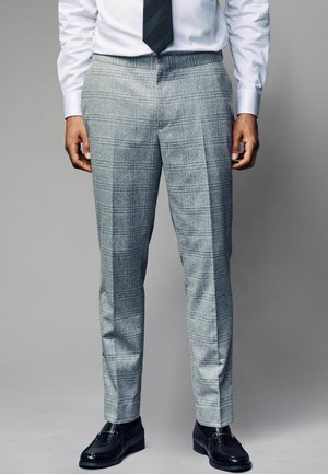 Pantalons à carreaux gris en tissu texturé, avec une coupe ajustée et un pli net, associés à des chaussures en cuir noires.
