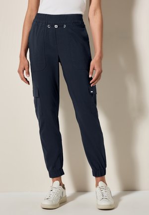 Kvinde iført navyblå cargo joggerbukser med elastiske manchetter og hvide sneakers, stående mod en ensfarvet beige baggrund.