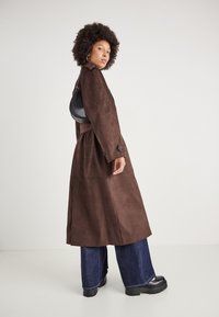 Cappotto trench in suede marrone con vita regolabile, grandi bottoni e spacchi laterali. Abbinato a jeans di denim a gamba larga e stivali neri chunky.