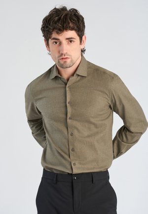 Man met krullend donker haar, die een olijfgroen geknoopt overhemd en zwarte broek draagt, staat met handen achter zijn rug.