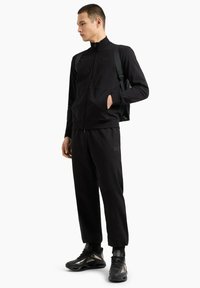 Completo sportivo nero con giacca con zip e pantaloni, realizzato in tessuto liscio, con un logo sulla giacca e sui pantaloni, abbinato a sneaker nere.