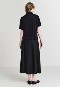 Donna che indossa una maglietta nera a maniche corte, una gonna midi nera, delle scarpe flat in tessuto nero e braccialetti beige, in piedi davanti a uno sfondo chiaro e uniforme.