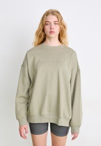 Olīvu zaļš oversized džemperis, kas izgatavots no mīksta kokvilnas, ar nenozīmīgu reljefu "adidas" logo. Apaļa kakla izgriezums un garas piedurknes.
