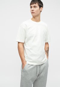 Λευκό βαμβακερό t-shirt με στρογγυλή λαιμόκοψη και κοντά μανίκια. Διαθέτει ένα μικρό, διακριτικό λογότυπο στο στήθος. Φορώντας ελαφριά γκρι φόρμες.