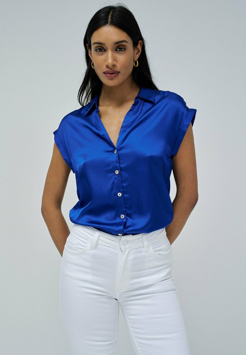 Camisa corta de satén azul con cuello y botones al frente, combinada con jeans blancos ajustados. Textura suave y contraste de color brillante.