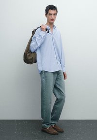 Ljusblå randig skjorta, löst sittande urtvättade jeans och bruna mockaloafers. Modellen bär en olivgrön tygväska.