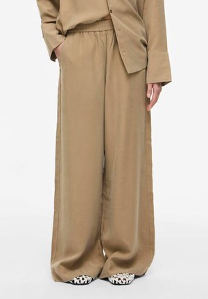 Osoba koja nosi široke bež hlače s elastičnim pojasom i bijele tkanine cipele na navlačenje, stoji s jednom rukom u džepu.