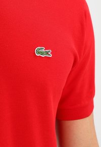 Rotes Polohemd mit strukturiertem Stoff. Ausgestattet mit einem grünen bestickten Krokodil-Logo auf der linken Brust. Kurze Ärmel und gerippter Kragen.