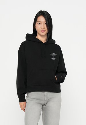 Svart hettegenser laget av bomull, med en frontlomme, snor i hetten og et grafisk trykk på venstre bryst. Kombinert med lys grå jeans.