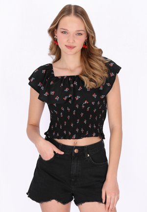 Junge Frau mit welligem blondem Haar trägt ein schwarzes, geblümtes Off-Shoulder-Oberteil und schwarze, hoch taillierte Jeansshorts mit ausgefranstem Saum.