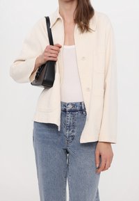 Jasna beżowa kurtka zapinana na guziki o luźnym kroju, z dwiema kieszeniami z przodu i kołnierzem, w połączeniu z niebieskimi dżinsami o wysokim stanie.