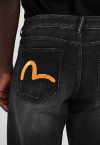 Jean en denim noir avec une broderie dorée sur la poche arrière. Le motif comprend un motif de lignes courbes. Décoloration subtile sur l'ensemble.