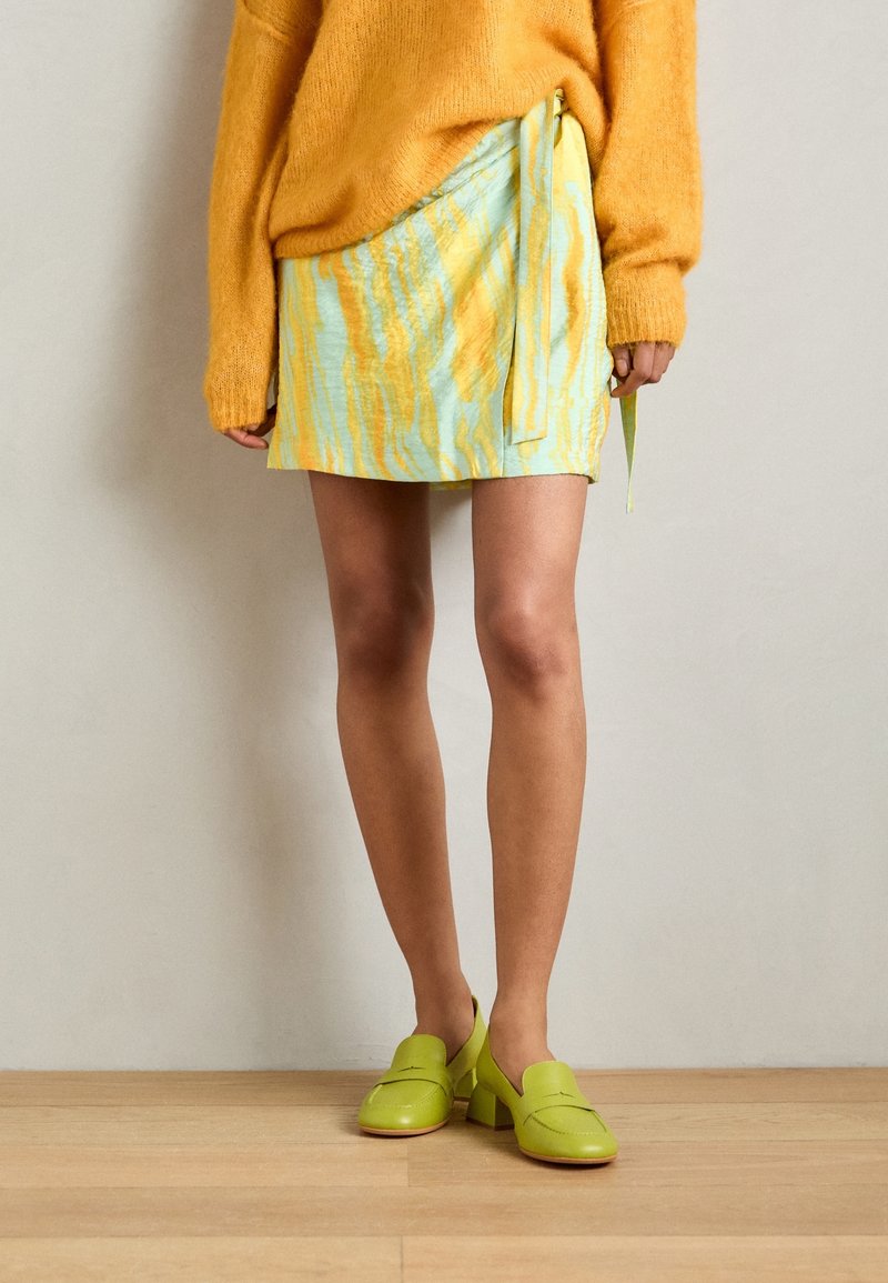 Falda azul clara con rayas abstractas amarillas, detalle de lazo lateral, combinada con zapatos de charol verde vibrante. El suéter es naranja y de corte oversized.