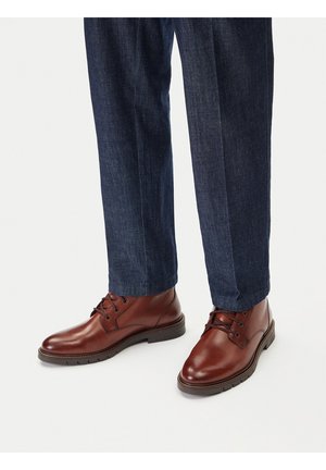 Brūnas ādas zābaki ar apaļām purngalām, auklu aizdari un teksturētu virsmu, kopā ar tumši zilām džinsa biksēm.