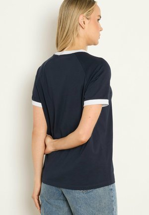 Vrouw met rug naar de kijker, gekleed in een marineblauw shirt met korte mouwen en witte bies, en een lichtblauwe spijkerbroek, staand tegen een effen achtergrond.