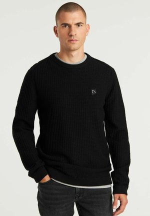Schwarzer Pullover mit strukturiertem Waffelstrickdesign, Rundhalsausschnitt und langen Ärmeln. Mit einem kleinen Logo-Patch auf der oberen linken Seite.