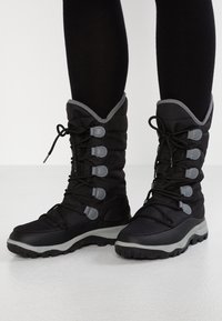 Bottes noires imperméables avec une tige rembourrée, des accents gris, des lacets et une semelle en caoutchouc texturé pour une meilleure adhérence. Hauteur mi-mollet, design fonctionnel.