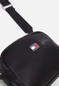 Bolso bandolera negro hecho de material sintético liso. Presenta una forma rectangular y un logo de Tommy Jeans en un cuadrado en la parte frontal.