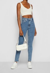 Blå denimhögmidjade jeans, krämfärgad ribbad crop top och vit handväska med logotyp. Vita sneakers fulländar outfiten.