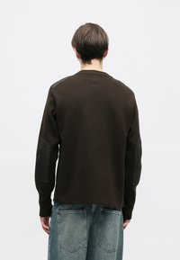 Barna pulóver kötött felülettel, fekete bőr váll- és könyökrátétekkel. Bő szabású, kerek nyakú és hosszú ujjú.