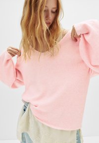 Svetlo roza oversized pulover z širokim dekoltejem in balonastimi rokavi, izdelan iz mehkega materiala, prekrit s svetlo, čipkastim zgornjim delom.