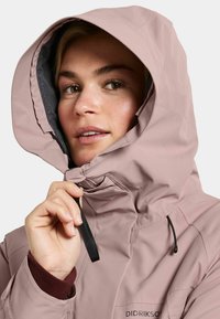 Chaqueta impermeable de color rosa claro con cuello alto y capucha, presenta un cierre de cremallera negra y cordón ajustable. Textura suave, diseño deportivo.