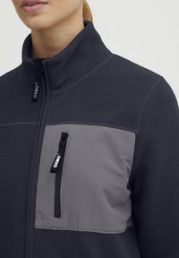 Fleece jacket in dark navy with a gray front panel. Features a high collar and zippered chest pocket with branded pull. Soft, textured material.