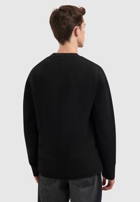 Zwarte gebreide trui met een ronde halslijn, geribde manchetten en zoom. Gelaagd stof. Model is van achteren afgebeeld, draagt donkere jeans.