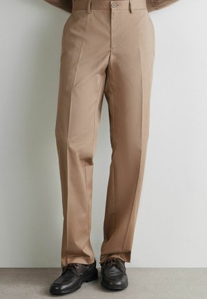 Beige bukser med lige ben og plisseringer foran, båret med sorte læder snørebåndssko, mod en ensfarvet lys baggrund.