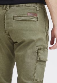 Olijfgroene cargobroek van duurzaam stof met een achterzak en een zijflapzak, voorzien van een leren merkembleem.