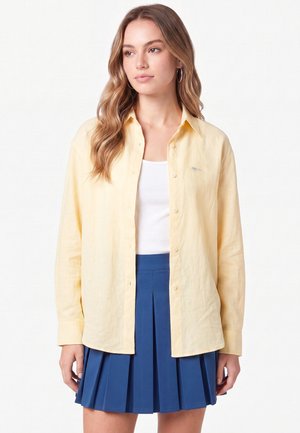 Jeune femme aux longs cheveux ondulés portant une chemise jaune clair à boutons par-dessus un top blanc et une jupe plissée bleue, debout devant un fond uni.