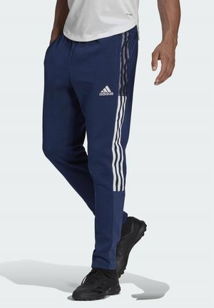 Mężczyzna ubrany w granatowe spodnie dresowe Adidas z białymi paskami i czarne trampki, stojący na jednolitym tle.