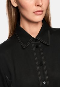 Schwarzes Hemd mit Kragen, kontrastierenden weißen Nähten und schwarzen Knöpfen. Der Stoff wirkt glatt mit leichtem Glanz.