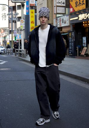 Junger Mann, der auf einer Stadtstraße geht, trägt eine gemusterte Mütze, eine schwarze Jacke, ein weißes Hemd, lockere schwarze Hosen und schwarz-weiße Turnschuhe.