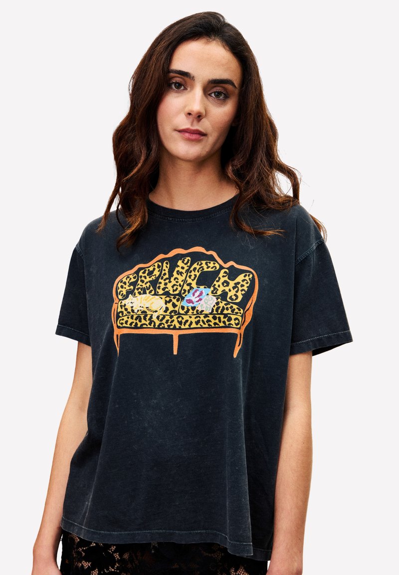Femme aux cheveux bruns ondulés portant un T-shirt noir avec un graphique de canapé à imprimé léopard et des coussins colorés.