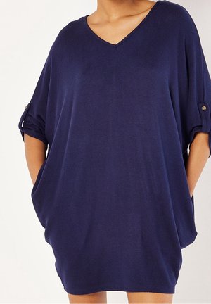 Vrouw draagt een losse marineblauwe jurk met V-hals, opgerolde mouwen bevestigd met knopen en handen in de zijzakken.