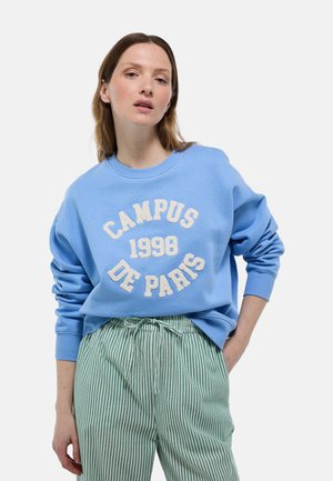 Pull bleu avec « CAMPUS 1998 DE PARIS » en lettres blanches texturées, avec un col rond et une coupe ample, associé à un pantalon rayé vert.