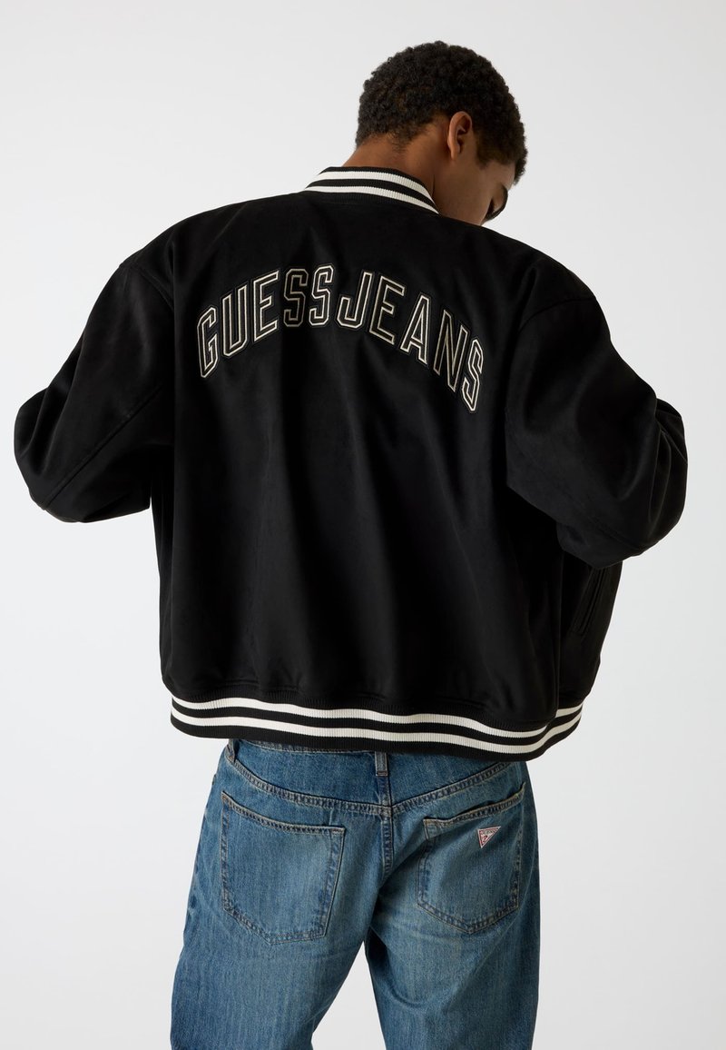 Giacca bomber nera con polsini e orlo a coste bianchi a strisce. "GUESS JEANS" ricamato in bianco sulla schiena. Texture liscia, vestibilità rilassata.