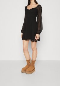 Svart klänning med texturerade genomskinliga ärmar, fyrkantig halsringning och utsvängd kjol. Parat med bruna plattformsskor med en kontrasterande sula.