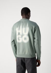 Zöld pulóver kerek nyakkal és hosszú ujjakkal. Nagy fehér "HUGO" felirat a hátsó részen, texturált szövet, bordázott mandzsetták és alsó szél.