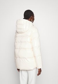 Vadderad vit pufferjacka med en glansig yta, en huva och en fyrkantig silhuett. Har bred horisontell quiltning och sänkta axlar.